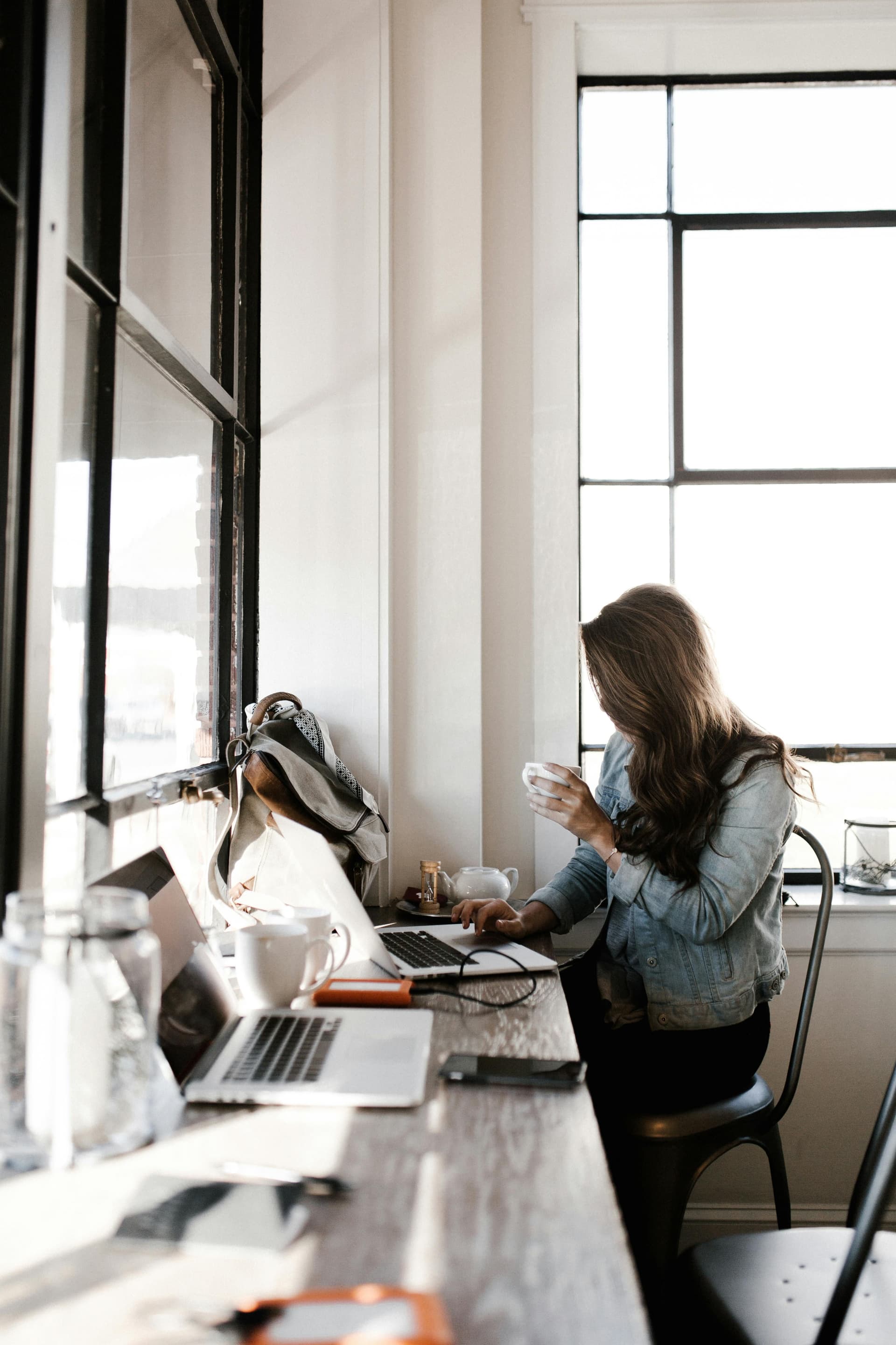 A person sits at a long wooden table near a large window, working on a laptop while holding a cup. Several laptops, notebooks, and cups are spread across the table, with natural light filling the space and creating a relaxed, café-like work environment.