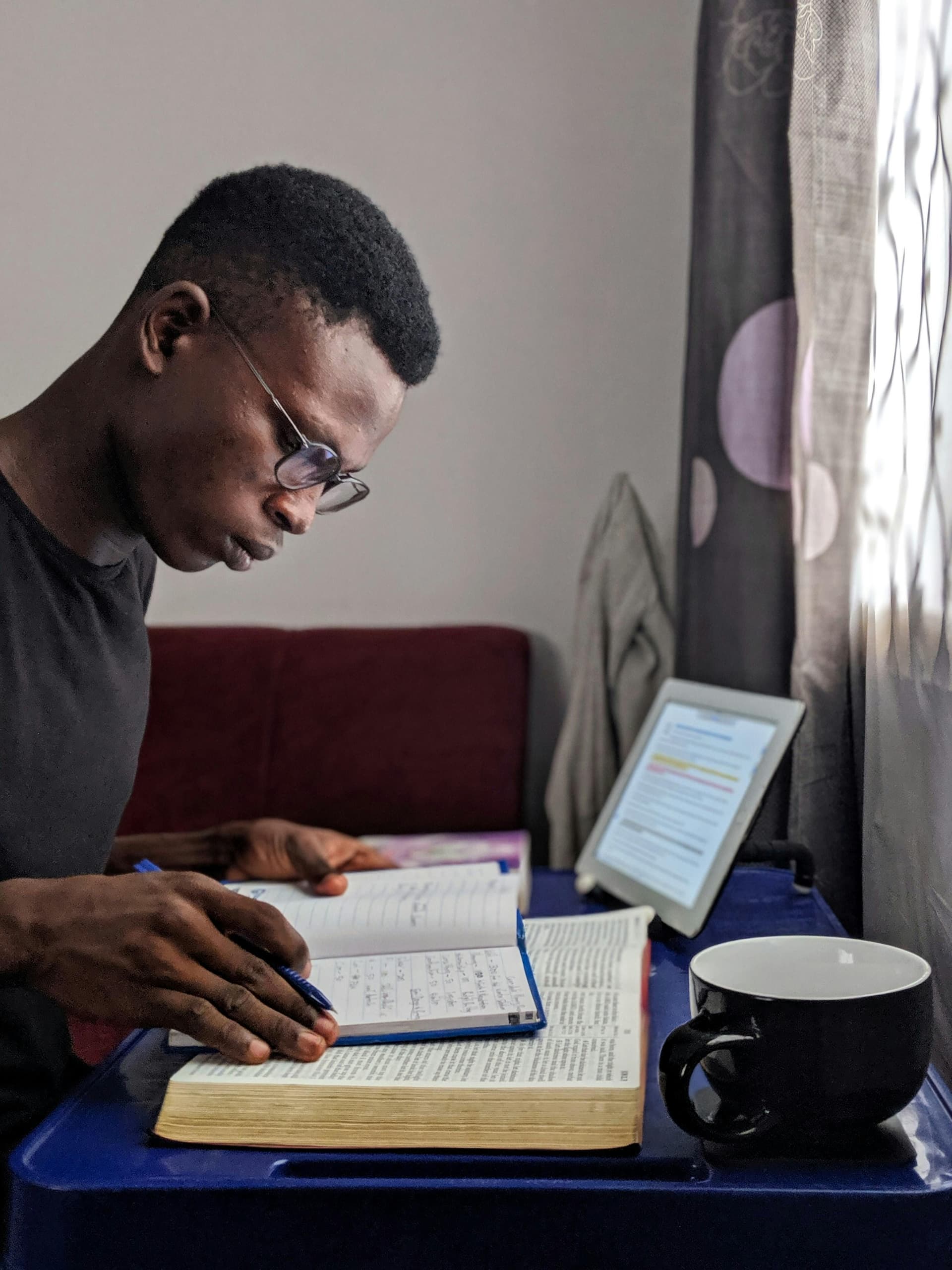 A person wearing glasses studies at a small table, writing notes in a notebook placed on top of an open book. A tablet displaying highlighted text and a black mug sit nearby by a window with sheer curtains, creating a quiet, focused study atmosphere.