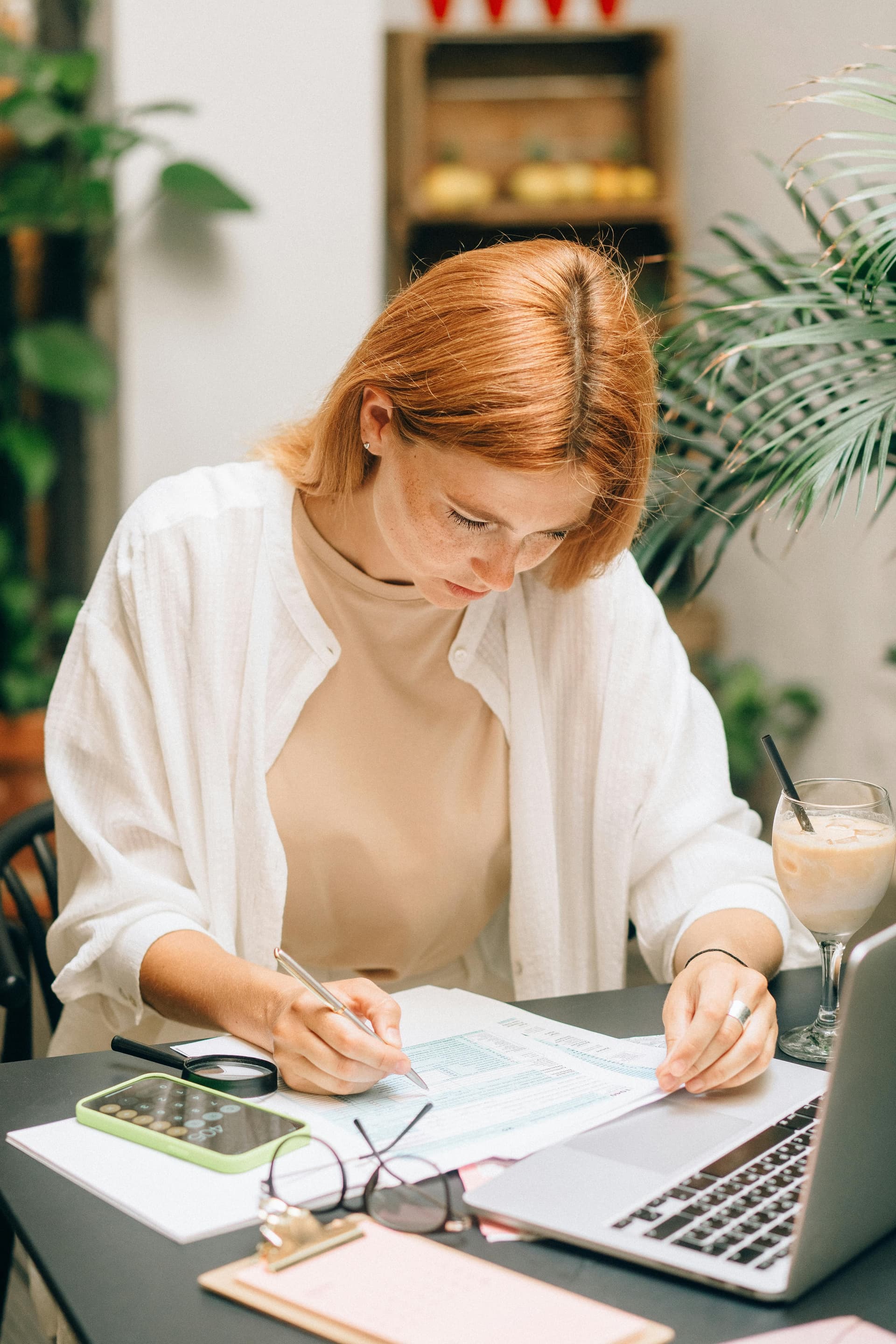 A person sits at a table reviewing printed documents and writing notes with a pen. A laptop, calculator, glasses, and a drink are arranged nearby, with plants in the background, suggesting a focused work or study moment in a bright, calm setting.