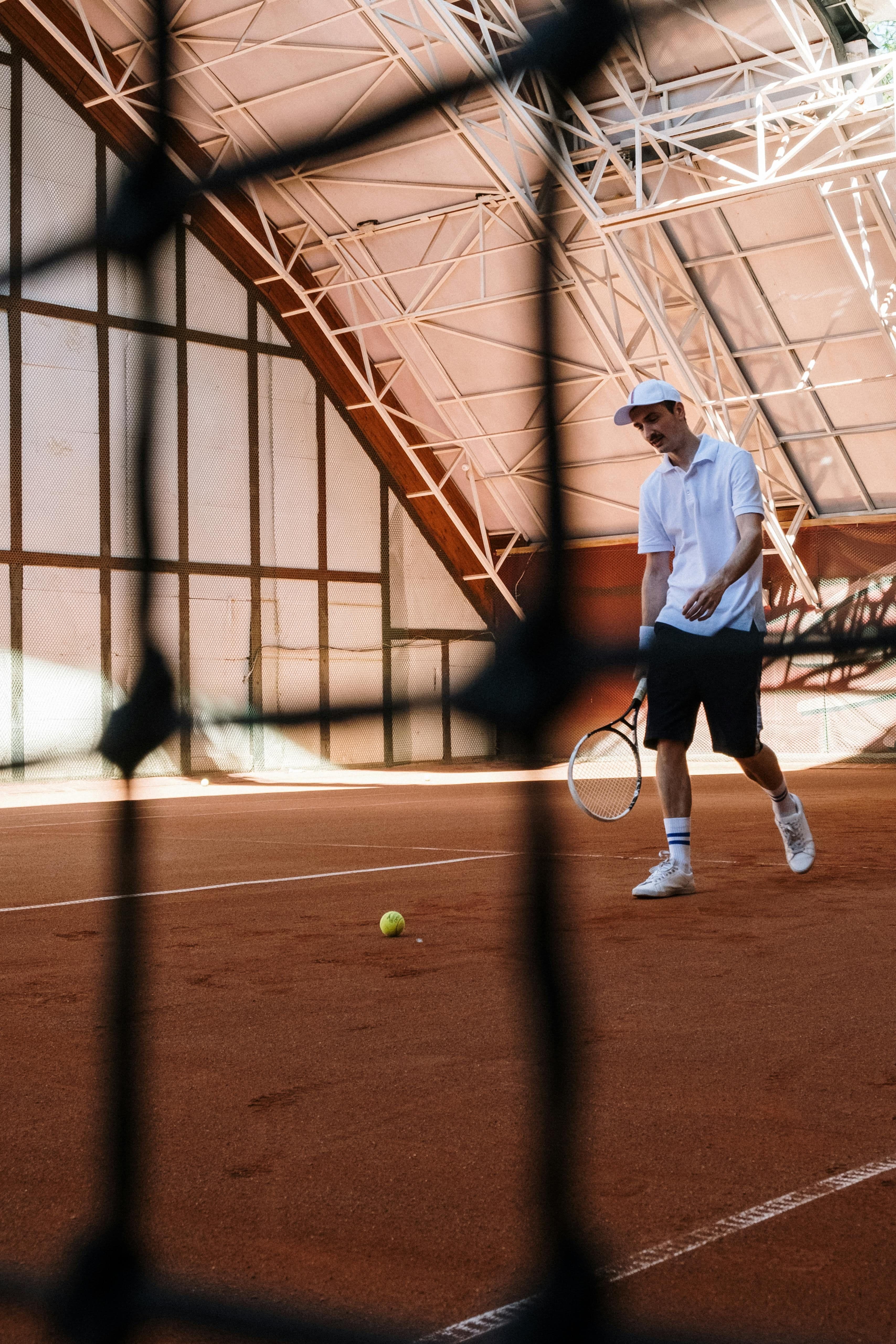 A tennis player walks toward a ball on an indoor clay court, holding a racket. The scene is viewed through the net, with the court’s metal roof structure visible above, conveying an active sports setting.